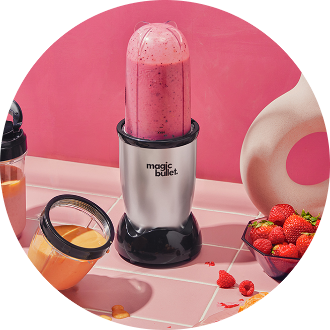 magic bullet and accessories filled with smoothie next to fruit against a pink background.